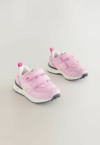 Rózsaszín sportcipő két tépőzáras pánttal, fehér foltos középtalppal, fekete gumi talppal és egy kis élénk rózsaszín húzófüllel a saroknál.