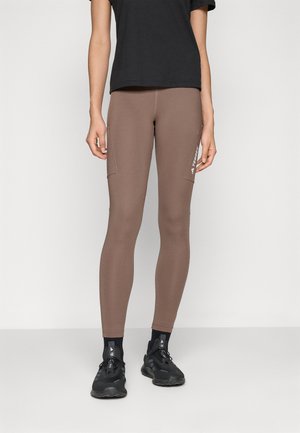Brune leggings med en tettsittende design, med sidelommer og en liten hvit logo. Outfiten er kombinert med svarte treningssko og en svart topp.
