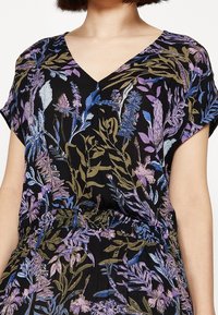 Femme portant une robe noire avec un motif floral bleu, violet et vert, décolleté en V, montrant le haut du torse et des manches courtes.