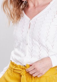 Bílý pletený kardigan s vzorem listů, s hlubokým výstřihem do V a knoflíky. V páru se žlutou sukní s vysokým pasem a uvázaným pásem.