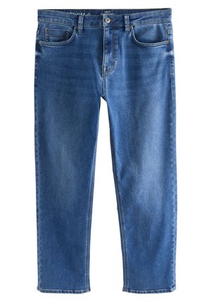 Person iført blå jeans med lige ben og brune ruskindssnøresko, stående mod en neutral baggrund.