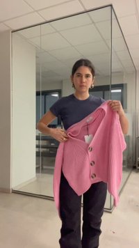 Frau hält einen pinkfarbenen, geknöpften Cardigan und steht in einem Raum mit Glaswänden und einer gefliesten Decke.
