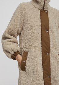 Beige fleecejacka med bruna tryckknappar framtill, texturerad tyg, sidofickor och ribbade ärmslut. Hög krage för extra värme.