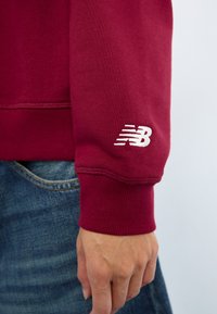Burgundijos spalvos megztinis su gipiūro rankovėmis, minkšta tekstūra ir balta „New Balance“ logotipu ant rankovės. Derinamas su mėlynomis džinsinėmis kelnėmis.