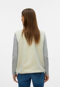 Gilet en polaire de couleur crème doux, sans manches avec un dos texturé, porté sur une chemise à manches longues gris clair, associé à un jean bleu.
