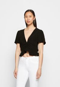 Top preto cropped com mangas curtas e largas, decote em V profundo, design frontal torcido e barra com babados. Combinado com jeans brancos.