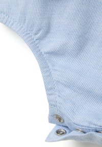 Tejido de mezclilla azul claro con una textura acanalada, que presenta broches de metal a lo largo del borde. Dobladillo curvado y un acabado suave visible.