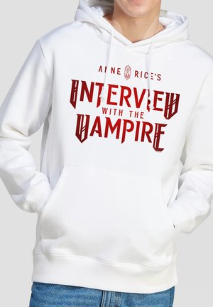 Person wearing a white hoodie with red text reading "Anne Rice's Interview with the Vampire" and hands in front pockets.