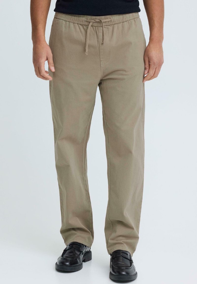 Homme portant un pantalon beige avec cordon de serrage et des chaussures en cuir noir, debout les mains détendues le long du corps, sur un fond clair uni.