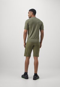 Mann, der mit dem Rücken steht, trägt ein olivgrünes kurzärmliges Hemd, passende Shorts, schwarze Socken und schwarze Turnschuhe mit grünen Sohlen.
