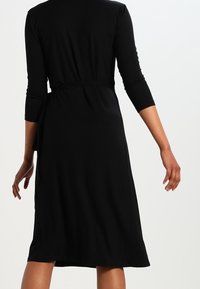 Svart ribbad klänning med trekvartsärmar och ett knytbälte i midjan. Kantlinjen faller nedanför knäet och har en slät textur.