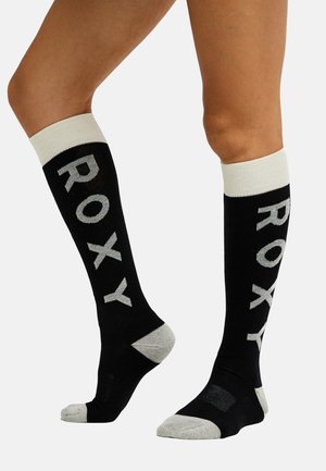 Jambes portant des chaussettes noires montantes aux genoux avec des bordures blanches, des orteils et talons gris, et un grand texte gris « ROXY » sur les côtés.