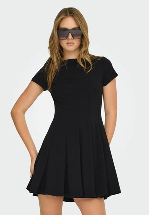 Kobieta o długich włosach, ubrana w duże czarne okulary przeciwsłoneczne i czarną plisowaną sukienkę z krótkim rękawem, pozująca z jedną ręką na biodrze.