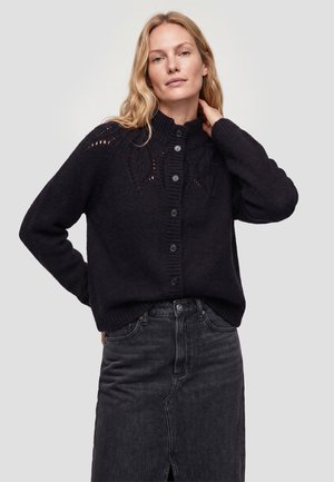 Kvinde med langt blondt hår iført en sort knaplukket striktrøje og mørke højtaljede jeans, stående med den ene hånd, der rører ved hendes nakke.