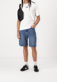 Bílá bavlněná tričko s logem, modré džíny šortky a černé tenisky s bílými pruhy. Černá křížová taška přes levé rameno.