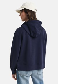 Marineblå hettegenser med stor hette, ribbestrikkede mansjetter og avslutning, kombinert med en hvit caps. Materialet ser mykt og texturert ut; avslappet passform.