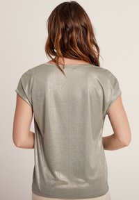 Top corto a maniche corte di colore verde metallico chiaro, con una texture liscia e una leggera lucentezza. Presenta maniche arrotolate e una vestibilità rilassata, senza motivi visibili.