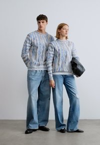 Δύο μοντέλα φορούν ταιριαστά ελαφριά μπλε πλεκτά πουλόβερ με μακριά μανίκια, συνδυασμένα με φαρδιά μπλε τζιν παντελόνια. Κρατείται μια μαύρη τσάντα.