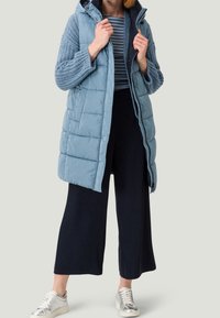 Light blue puffer vest with a hood, quilted texture, and ribbed sleeves. Paired with navy wide-leg trousers and silver sneakers.