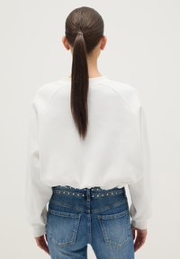 Sudadera blanca recortada con mangas raglán, combinada con jeans azules de talle alto que tienen una cintura con tachuelas. Textura de tela suave.