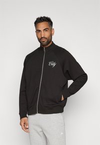 Άντρας με κοντά σκούρα μαλλιά και μούσι, φορώντας μαύρο φούτερ με φερμουάρ με το κείμενο "City" και ανοιχτό γκρι φόρμες, που στέκεται μπροστά σε απλό φόντο.