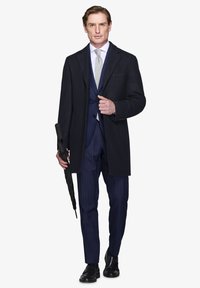 Skræddersyet mørk frakke over en navy stribet jakkesæt, parret med et lyst slips og sorte sko. Holder en sort paraply.