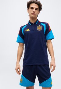 Young man wearing a navy blue and light blue Spain soccer jersey and matching shorts with red and yellow accents.