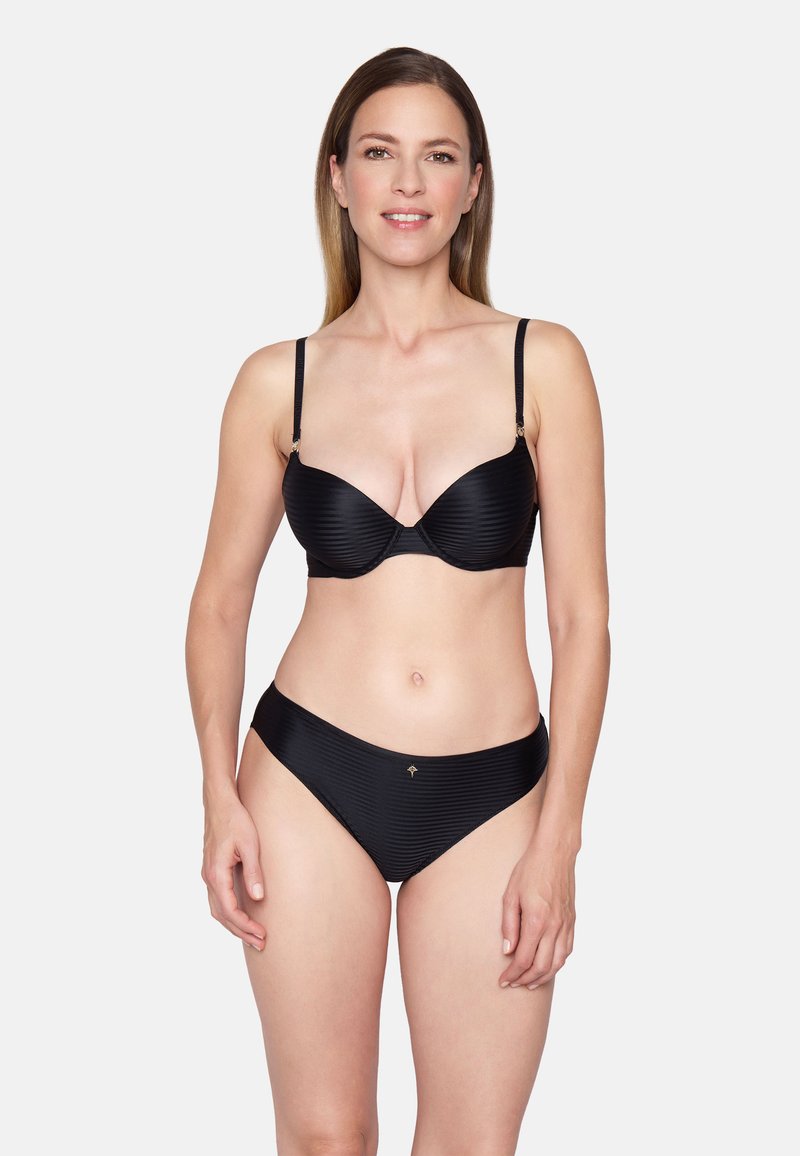 Femme souriante aux cheveux raides portant un soutien-gorge noir rayé et une culotte assortie, debout devant un fond blanc uni.