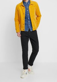 Geel corduroy jack over een blauw denim overhemd, gecombineerd met zwarte broek en witte sneakers met rood- en blauwachtige strepen.