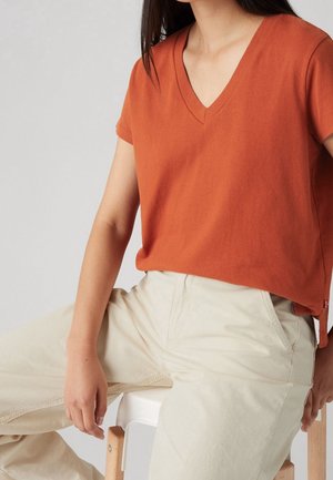 Personne portant un t-shirt orange à manches courtes en V et un pantalon beige clair, assise sur une chaise en bois devant un fond uni.