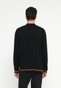 Fekete kötött pulóver, függőleges csíkokkal, bordázott gallérral és narancssárga díszítéssel az alján. Hosszú ujjú, laza szabású.