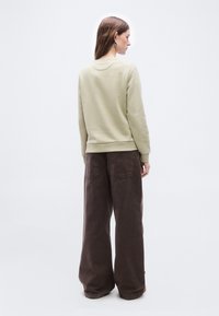 Γυναίκα με μακριά καστανά μαλλιά φοράει μπεζ φούτερ και φαρδιά σκούρα καφέ παντελόνια, στέκεται αντικρίζοντας ένα απλό φωτεινό φόντο.