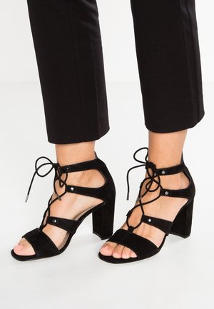 Zwarte suède hoge hakken met open teen, enkelbandjes en vetersluiting met ronde hardware-accenten. Vierkante hak.