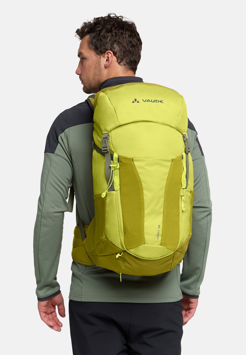 Ein Mann trägt eine grün-graue Jacke und schwarze Hosen und hat einen leuchtend gelb-grünen Vaude-Rucksack mit mehreren Fächern auf dem Rücken.