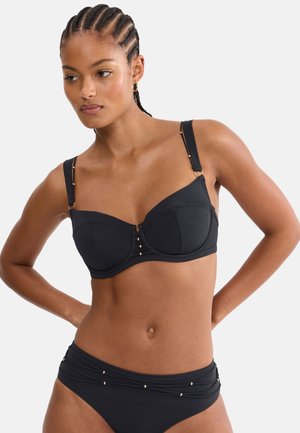 Frau mit geflochtenem Haar modelliert einen schwarzen Bikini mit goldenen Akzenten, Hände auf Hüfte gestützt, vor einfachem hellem Hintergrund.
