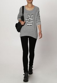 Pull gris avec un motif graphique de la main « paix » en noir et blanc à rayures, associé à un jean skinny noir et des bottines noires à clous.
