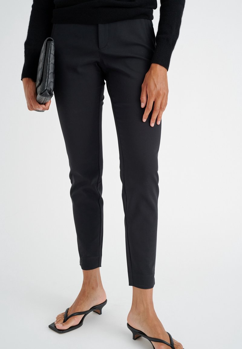 Personne portant un pantalon noir slim, des sandales à talons noires, tenant une pochette noire, debout sur un fond blanc.