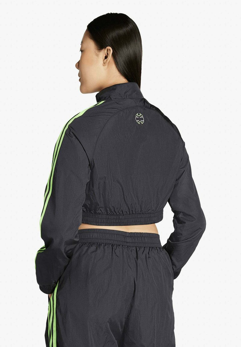 Kvinna som står med ryggen mot och bär en svart kort träningsjacka och matchande byxor med neongröna ränder längs ärmar och sidor.