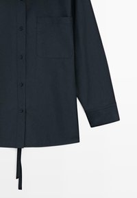 Ciemnogranatowa koszula na długi rękaw zapinana na guziki z kieszenią na piersi oraz materiałowymi troczkami zwisającymi z dołu na białym tle.