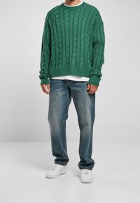Pull vert à maille torsadée en tissu texturé, associé à un jean bleu délavé et des baskets blanches. Le pull présente un col rond et des poignets côtelés.