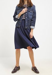 Jeansjacka med knäppning fram, vikta ärmar; blommig topp; plisserad marinblå kjol; bruna läderskor med snörning och blank yta.