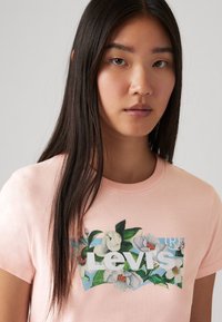 T-shirt en coton rose clair avec un graphique floral et le texte "Levi's" en blanc. Manches courtes et design à col rond. Texture lisse.