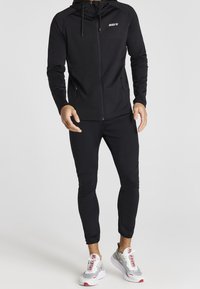 Svart zip-up hoodie med huva och logotyp; tillsammans med svarta slim-fit byxor. Skorna är grå och vita sneakers med röda accenter.