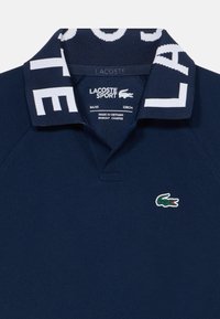 Polo marine avec un col imprimé de logo blanc, petit emblème de crocodile sur la poitrine et une texture lisse. Fabriqué en tissu respirant.
