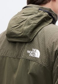 Άνδρας που φοράει μπουφάν The North Face σε ελαιόπρασινο χρώμα με κουκούλα, ορατό λογότυπο στο πάνω μέρος της πλάτης και λεπτομέρεια με σχέδιο στο ύφασμα στους ώμους.