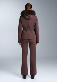 Tamnoljubičasta gornja odjeća s kapuljačom obloženom krznom, pojasom u struku i prilagođenim krojem. Hlače na zvono imaju detalje s patentnim zatvaračem oko gležnjeva. Glatki, rastezljivi materijal.