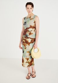 Blomsterprydet kjole til kneet i myk stoff, med nyanser av grønn, brun og beige. Kombinert med en vevd gul håndveske og brune sandaler.