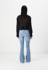 Femme aux longs cheveux foncés portant un haut noir à manches longues et un jean évasé bleu clair, debout de dos sur un fond blanc.