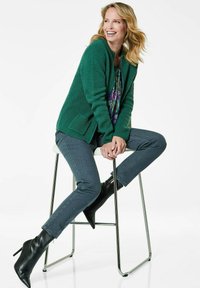 Grüne strukturierte Jacke mit zwei Taschen, kombiniert mit einem gemusterten Oberteil, grauen Skinny-Jeans und schwarzen spitzen Stiefeln, sitzt auf einem Hocker.