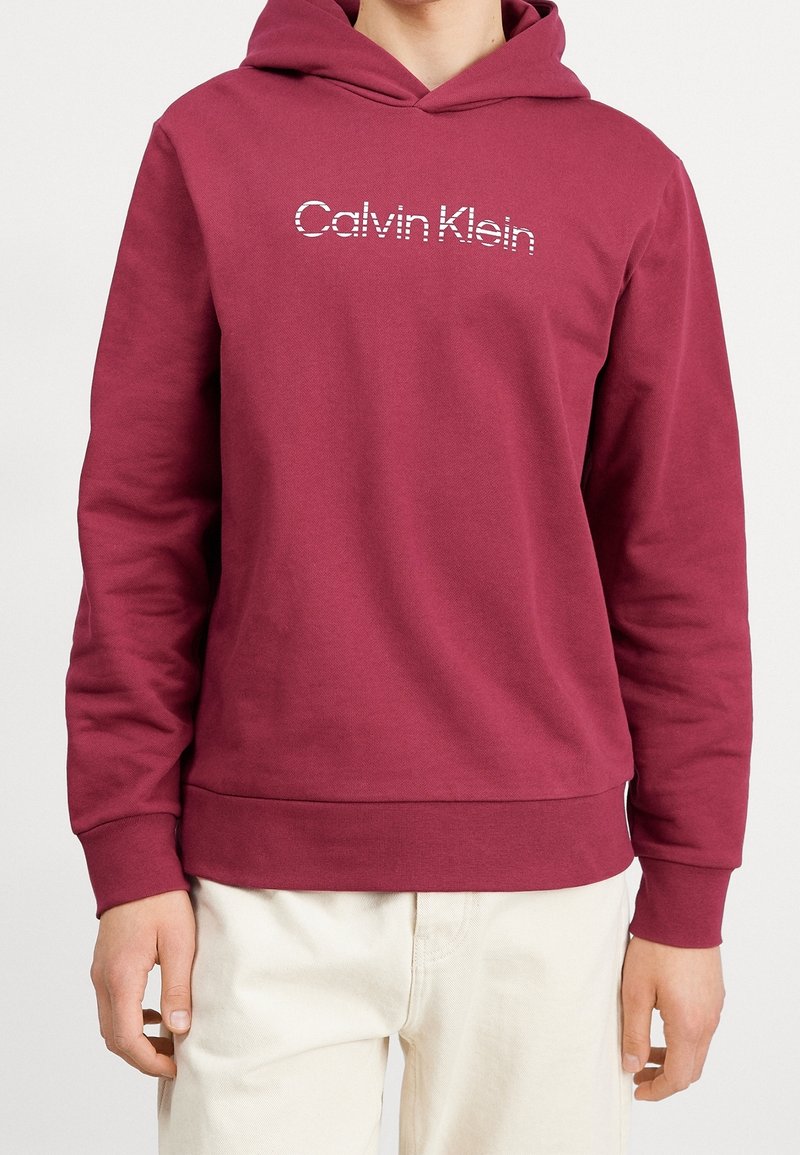 Maroonowy sweatshirt z kapturem, wykonany z miękkiego materiału, z prążkowanymi mankietami. Posiada biały logo "Calvin Klein" na klatce piersiowej. Prosty design.
